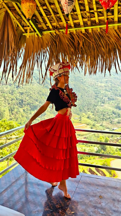 A woman in traditional attire stands gracefully on an elevated outdoor platform with a thatched roof, overlooking a vast mountainous landscape in Thông Nguyên Commune, Vietnam. She is barefoot, wearing a black crop top, a vibrant red, multi-tiered skirt, and an elaborate red and silver headdress. Decorative elements matching the headdress are also visible on her chest and shoulders. Her right hand delicately holds a section of her skirt, while her left hand rests on her hip, suggesting a pose of movement or dance.

Above her, the roof structure is made of dried palm fronds and bamboo supports, from which two colorful lanterns hang: one yellow with butterfly motifs and one white with floral patterns, both featuring tassels. The background is dominated by lush green hills and valleys, characteristic of the Vietnamese countryside, with hints of terraced fields visible in the distance. A dark railing runs along the edge of the platform.

The floor beneath her is dark and reflective, appearing to be plywood. Visible text on the floor includes phrases like "TRACHOS PLYWOOD," "MOOD TRACHDUY PLYWOOD TRACK," "NA PREN," "QUY PLYWOODTRA," and "PREMILL," suggesting product branding. The scene is brightly lit by natural daylight, casting shadows on the reflective surface, indicating a clear and sunny day. The overall impression is a cultural portrait set against a stunning natural backdrop.
