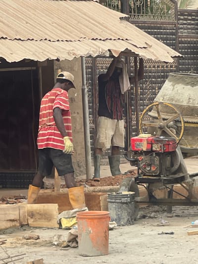 Two men work on a construction site. The man in the foreground wears a red and white striped shirt, black shorts, and a white hat. He also wears yellow gloves. A second man, standing behind the first, wears a red and black plaid shirt. A cement mixer with a yellow wheel sits in the background. An orange bucket and other construction materials are visible. The ground appears to be dirt or concrete. Corrugated metal forms a partial roof.