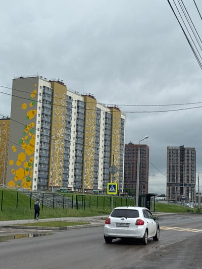 This image captures an urban street scene in Kondratovo, Russia, under an overcast sky. The foreground is dominated by a modern, multi-story residential building on the left, featuring a distinctive facade with large sections of yellow paint adorned with white and green hexagonal patterns. Further in the background, two taller, darker high-rise buildings are visible, one of which prominently displays the text "ИНГРУПП" (INGROUP) vertically on its side.
On the right side of the frame, an asphalt road features a clearly marked pedestrian crosswalk. A white Toyota car, with the Russian license plate "О384ХС 159," is positioned on the road near the crossing. A bright yellow pedestrian crossing sign stands prominently beside the road. Between the road and the main residential building, a grassy strip with a fence is visible, where a person in dark clothing, possibly wearing a face mask, is walking. A bus stop shelter is also partially visible further down the road.
Overhead, several power lines stretch across the light gray, cloudy sky. A street light is also part of the urban infrastructure visible in the scene. The road appears slightly wet, suggesting recent rain or damp conditions. The scene depicts a typical daytime setting in a developed residential area.