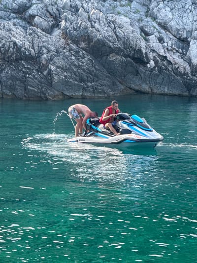 This media file shows two people on a jet ski on a sunny day, near a rocky coastline in Dubraljevina, Montenegro. The water is a clear, turquoise green, with sunlight reflecting off its surface. The rocky cliff face is gray and textured, showing signs of erosion.
The jet ski is white and blue, with the number "BD 1390" visible on its side, along with the "Yamaha" logo. One person, wearing a red life vest, is seated at the front of the jet ski, holding onto the handlebars. Another person, shirtless and wearing patterned swim shorts, is leaning back on the jet ski, with one leg extended and kicking water. They appear to be enjoying a recreational activity. The sunlight suggests it is daytime, likely afternoon given the shadows and the bright water. The overall mood is one of leisure and fun.
