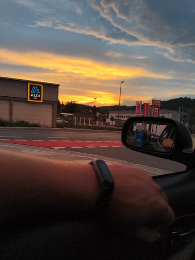 The image depicts a view from inside a vehicle looking outwards at a street scene during sunset in Pegnitz, Germany. The sky is illuminated by a vibrant orange and yellow sunset, with scattered clouds. On the left side of the frame, the ALDI Süd store is visible with its distinctive logo. Further down the street, a REWE CENTER store is identifiable by its red and white banners. A car's side mirror reflects a blue object labeled "JARA". The foreground shows a person's arm with a fitness tracker on their wrist, resting on the car door. A red painted crosswalk is prominent on the road. The overall environment suggests a commercial area within a town or city.