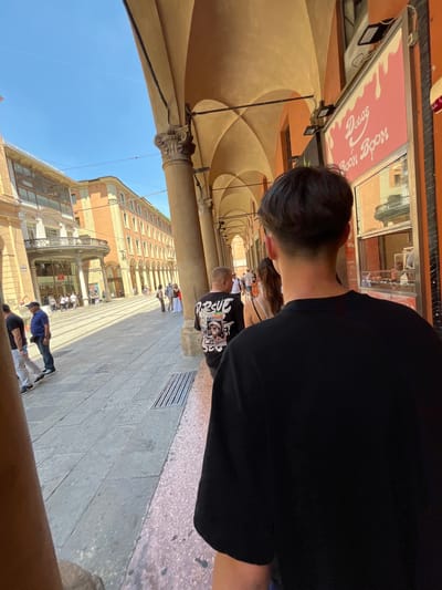 The image is taken under the arcades of Bologna, Italy, which are characterized by tall, arched structures supported by columns. These arcades line the streets and buildings. In the foreground is the back of a person wearing a black shirt. Along the street, there are pedestrians walking, and a shop identified as "H&M" is visible. The sky is clear and blue. A sign for "Dolce Spoon" is partially visible. The image appears to be taken by a person looking along the arcade. The presence of the arcades and the style of the buildings are characteristic of Bologna.