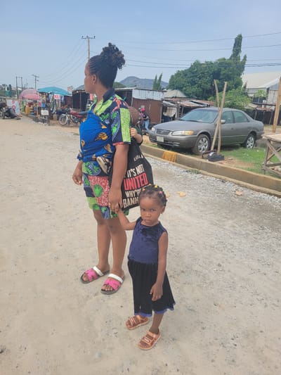 The image depicts a woman and a young girl standing on a dirt road in Kwamba, Nigeria. The woman, with braided hair, is wearing a colorful patterned dress and pink sandals. She is carrying a child on her back, and the child wears a black t-shirt with white writing. The young girl standing beside her is wearing a dark blue dress and brown sandals. In the background, there is a car parked next to a curb and multiple street vendors.