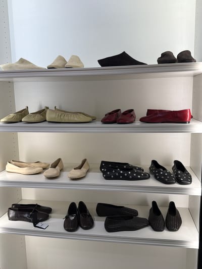 The image displays a closet with multiple shelves organized to store shoes. The closet appears to be part of a retail store or a well-organized personal wardrobe. The lighting suggests it is daytime, with artificial light illuminating the interior of the closet.
On the leftmost shelf, there are several dark-colored, soft-brimmed hats, stacked on top of each other. Below the hats, a pair of black boots are displayed, with one boot positioned upright and the other lying on its side.
The second shelf from the left holds a pair of red leather ankle boots. Below these, a pair of darker red leather shoes with buckles are visible.
The third shelf features black slip-on shoes adorned with silver polka dots. There are two pairs of these shoes, with the top pair facing forward and the bottom pair positioned slightly behind.
On the fourth shelf from the left, there are two pairs of dark brown woven slip-on shoes, stacked one above the other. Below these, two pairs of dark brown leather loafers are displayed, one of which has a tag attached.
The fifth shelf contains two pairs of light beige ballet flats.
The shelf furthest to the right showcases several pairs of light beige shoes. The top two pairs are slip-on shoes, with one pair facing forward and the other angled. Below these, two pairs of ballet flats are displayed.
The overall scene is neat and orderly, showcasing a variety of footwear organized by style and color. There is no indication of people or any specific activity taking place beyond the display of the shoes. The setting is an indoor closet, likely in a location with organized storage solutions. No visible text beyond a shoe tag on the bottom right shelf.