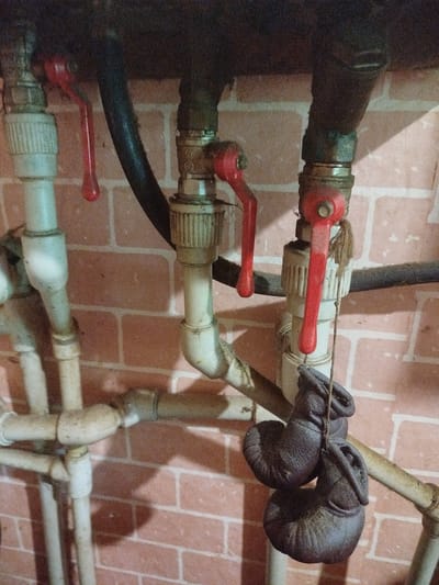 The image shows a close-up of plumbing pipes and valves mounted on a brick wall. Several red ball valves are visible, some in the open position. A black hose runs horizontally behind the valves. Below the valves, a pair of small, dark-colored objects, resembling miniature boxing gloves, hang from a string. The overall scene appears to be in an indoor utility area, likely a basement or service room, in the city of Kamianske, Ukraine. The lighting is dim, casting shadows on the wall.