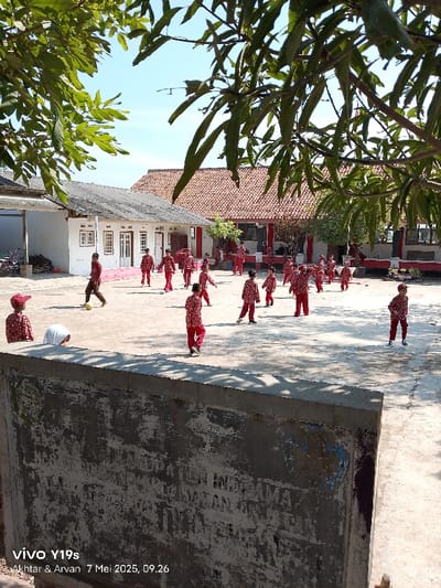 The image shows a schoolyard in Kecamatan Haurgeulis, Indonesia (latitude: -6.419, longitude: 107.972). A group of children, all wearing red shirts and pants, are playing in a large, paved courtyard surrounded by buildings. There are three main buildings with tiled roofs, one of which appears to be a school building with windows. The courtyard is partially shaded by a large tree in the foreground, whose branches are reaching out towards the buildings. The children are engaged in various activities. Some are running around, while others are kicking a soccer ball, which is visible in the center of the image. The sun is shining, and there is a cast shadow from the tree in the foreground. The ground is dusty and covered with some leaves and pebbles. A large, weathered concrete wall with faded writing on its surface is in the foreground. The wall is covered in a thin layer of dust and debris. The writing on the wall is obscured but is partially visible. The image appears to have been captured by a cell phone camera.