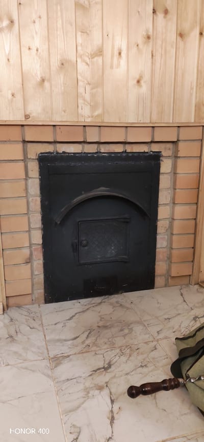The image shows a fireplace in Беркуты, Russia, with a black metal door set within a brick surround. The fireplace is located against a wall covered with light-colored wood paneling above the brickwork. Below the fireplace is a tiled floor with a marbled pattern. To the right of the fireplace, on the floor, is a bag or piece of material with what appears to be a wooden-handled barbecue skewer or tool resting on it. The bottom left corner of the image shows the text "HONOR 400".