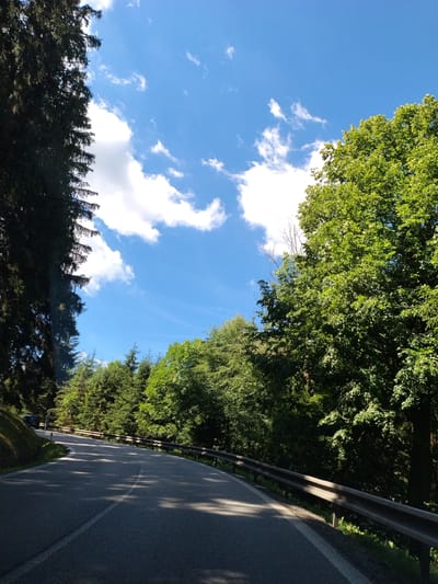 The image depicts a rural road winding through a dense forest on a sunny day. The road is paved and has a white line marking the center. A metal guardrail runs along the right side of the road. Tall, dark green coniferous trees line the left side of the road, while lush green deciduous trees form a thick canopy on the right. The sky is a vibrant blue with scattered white clouds. In the distance, a dark-colored vehicle is visible on the left side of the road, navigating the curve. The scene suggests a serene and natural environment, characteristic of a mountainous or forested region. While the city of Horní Vltavice, Czechia is mentioned, the image itself does not contain any specific landmarks or identifying features that would confirm its precise location within the city. The overall impression is one of a scenic drive through unspoiled nature.
