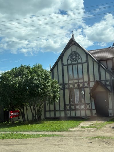 The image depicts a two-story residential structure of distinct Tudor Revival architectural style, featuring exposed dark timber framing over light-colored infill panels. The house is situated in what appears to be a semi-rural or suburban environment, potentially in Кварса, Russia. The front facade presents a prominent gable end, centrally adorned with a large, intricately carved round window (resembling a rose window) above multiple vertical window panes. Below these, a decorative horizontal band of quatrefoil-like patterns is incorporated into the timberwork. A recessed entrance with a small gabled porch and a dark door is visible on the lower right side of the facade, accompanied by a wall lantern. The house is topped with a steeply pitched roof, transitioning to a corrugated metal roof section on the right, from which a metal chimney or vent pipe emerges. A small finial crowns the main gable's apex.

To the left of the house, a large deciduous tree with dense green foliage partially obscures the building. Beneath this tree, a red rectangular sign with white Cyrillic text, "САЖЕНЦЫ" (SAPLINGS), is affixed to a dark fence, pointing towards the right. The ground in the foreground consists of a dirt or gravel path/road, transitioning into a strip of green grass containing some yellow flora, possibly dandelions. A lighter-colored, possibly concrete path leads from the foreground towards the house's entrance.

The sky above is bright blue with scattered, fluffy white cumulus clouds. Multiple parallel utility lines traverse the upper portion of the frame, horizontally spanning across the sky and above the house. No individuals are present within the scene. The lighting suggests a clear, sunny day, likely during late morning or early afternoon hours.