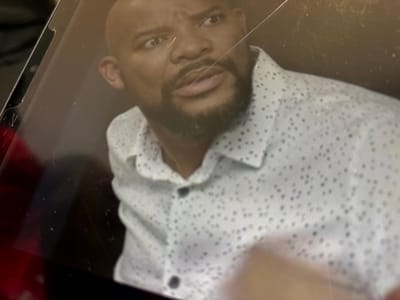 The image displays a photograph of a digital screen depicting an adult dark-skinned male with a shaved head and a dark beard. The man is wearing a light-colored, possibly white or light grey, collared shirt adorned with numerous small dark dots. His eyes are wide and focused slightly upward and to the viewer's left, with furrowed eyebrows, and his mouth is slightly agape, conveying an expression of surprise or concern. The screen itself exhibits multiple visible cracks or scratches, predominantly diagonal, across its surface, obscuring parts of the man's face and torso. A dark bezel or frame of the digital device is visible along the left edge of the image, showing minor damage or wear near the top left corner. A reddish reflection is present in the lower-left corner of the screen. The scene is located in Midrand, South Africa.