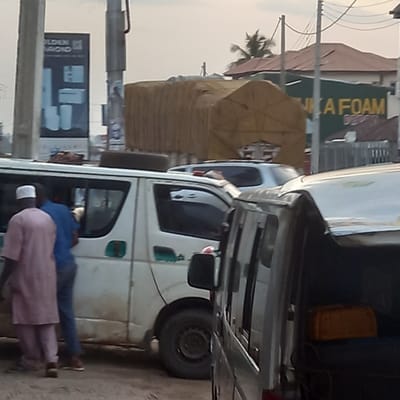 Two men stand next to a white van parked on a street in Dadi Riba, Nigeria. One man wears a pink shirt and white cap; the other, a blue shirt and pants. The van is loaded with goods covered with a brown tarp. A sign for "Foam" is visible in the background. A billboard showing bathroom fixtures stands on a pole.