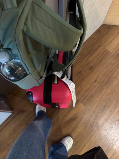 The scene depicts an overhead view of luggage and a person standing on a wooden floor. A green backpack filled with a water bottle is positioned near a red rolling suitcase with a baggage tag. The person is wearing blue jeans and white sneakers. The location is not identifiable as Bogota, Colombia.