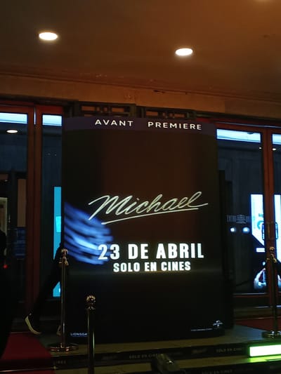 The image shows a promotional display for the movie "Michael" during an avant premiere at a cinema. The display is black with the title "Michael" in a cursive font, followed by "23 DE ABRIL" and "SOLO EN CINES" in white text. "AVANT" and "PREMIERE" are printed in smaller white letters near the top of the display. Gold-colored stanchions with ropes are visible on either side of the display, suggesting a red-carpet event or queue. A person wearing sneakers and black pants is walking behind the barrier. The text "LIONSGA", likely a production company, is visible near the bottom of the display. The background includes reflections and glimpses of the cinema's interior. Logos related to "SOLO EN CINES" are visible at the base of the display and on what seems to be the floor. "Universal Pictures" also appears on the display.