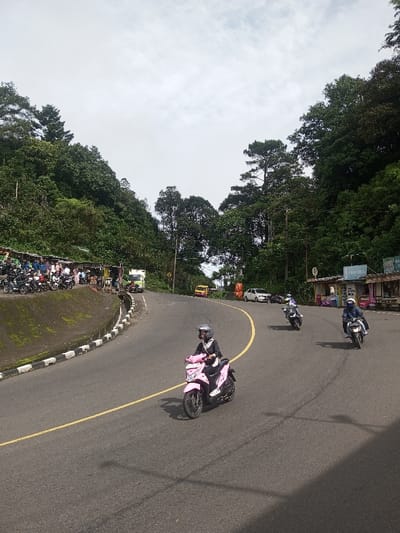 This image depicts a curved asphalt road traversing a heavily vegetated, hilly landscape identified as Sandamal Eliya, Sri Lanka, under an overcast sky during daylight hours. The road features a single solid yellow center line and a black and white striped curb delineating its left edge.
On the left side of the road, a raised earthen bank with green foliage leads to a series of approximately five informal roadside stalls or structures, characterized by various roofing materials. An estimated 15-20 motorcycles are parked in front of these stalls, where approximately 10-15 individuals are gathered, some standing, some seated. Further along this side, a light-colored box truck is visible.
On the right side of the road, dense trees cover an uphill slope. A building or larger stall with a partially visible blue sign, indicating "PKU CELL", is present. Multiple utility poles and power lines extend along this side. Beyond this point, a yellow utility vehicle and a white sedan are parked on the shoulder.
Three individuals are actively operating motorcycles on the road, all moving away from the viewer. In the foreground, a person wearing a black jacket, dark pants, and a gray full-face helmet rides a pink scooter/motorcycle on the left side of the yellow line. In the midground, two additional individuals are on motorcycles on the right side of the road; the first wears a light-colored helmet and jacket on a dark motorcycle, and the second wears a dark blue helmet and jacket on a dark motorcycle. Diffused light from the overcast sky casts subtle shadows on the road surface.