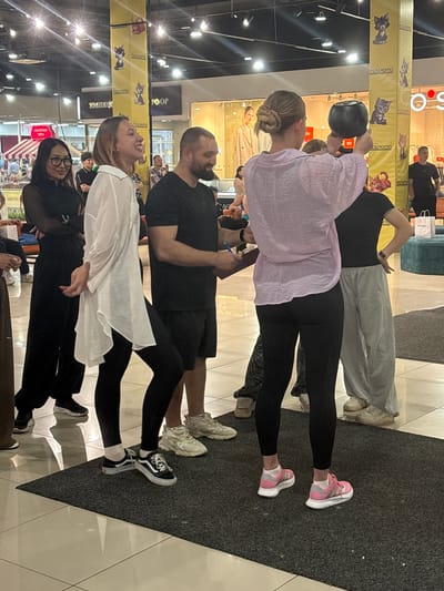 This image depicts an indoor shopping mall setting, likely in the city of Tri Protoka, Russia. In the foreground, a woman with blonde hair pulled into a bun is holding a dark, round object, possibly a kettlebell, in front of her. She is wearing a light pink, oversized shirt and black leggings. To her left, a man in a black t-shirt and black shorts is holding a clipboard and a pen, seemingly interacting with her. To the left of the man, a woman with dark hair and glasses, dressed in a black sheer top and black pants, stands looking to her right. Another woman in a white, flowing shirt and black leggings is positioned between the woman in glasses and the man. Behind the main group, other individuals are visible in the background, along with store displays and pillars adorned with images of cats. The floor is tiled, and a dark mat is laid out in the foreground. The overall atmosphere suggests a public event or promotion within the mall.