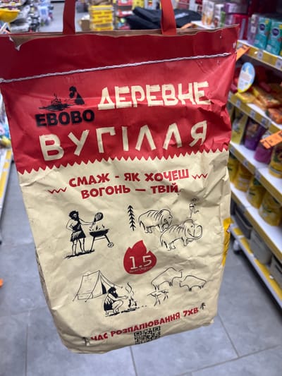 The image shows a bag of charcoal with a red and beige design. The bag features stylized drawings of people, animals resembling mammoths, and a campfire. Text in a Cyrillic script is visible on the bag, with large lettering in white and yellow. The charcoal bag is held by its red handles. In the background, there are shelves stocked with various products, typical of a retail store environment. The lighting suggests an indoor setting with artificial lights. The floor appears to be tiled. The overall scene depicts a product being presented in a commercial setting, likely a grocery or hardware store. There is no discernible indication of the specific location being Ivanivka, Ukraine, beyond the script on the bag which is consistent with languages spoken in that region.