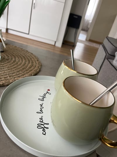 The image shows a light green, double-cup coffee mug with gold trim and gold handles resting on a white saucer. The saucer has black script that reads "coffee, love, bakery" with a small red heart. Two silver spoons are placed inside the cups. The background includes a section of grey upholstered furniture, a tan woven circular placemat, and a white kitchen cabinet. The location is not discernible from the image.