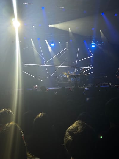 The image shows a concert performance at night. In the foreground, a crowd of people are visible, silhouetted against the stage lights. Some people are holding up their phones, presumably to record the event. The stage is brightly lit with various spotlights and LED panels, casting beams of white and blue light. On stage, musicians can be seen performing. One person is playing a keyboard, another appears to be a drummer, and a third individual is standing near a microphone. The background is dark, with some architectural elements of the venue possibly visible. The overall atmosphere suggests a lively and energetic concert. There is no information in the image to suggest this is in Palilula Urban Municipality, Serbia.