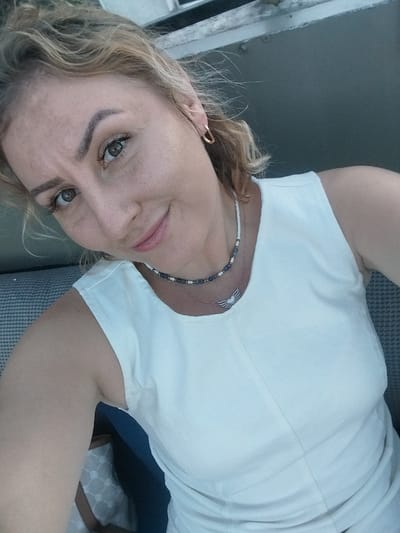 A medium shot portrait of a fair-skinned woman with blonde hair, looking towards the camera. She is wearing a white sleeveless top, a beaded necklace, and a hoop earring. Her head is tilted slightly to her left, and she has a subtle smile. The background is blurred, showing a dark gray wall or surface and the texture of a seat. No specific location in Istanbul, Turkey is identifiable from the image.