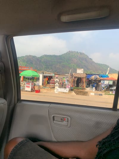 The image, taken from inside a vehicle, captures a roadside scene in Dutsen Alhaji, Nigeria. In the foreground, the interior of the vehicle is visible, including a portion of a person's leg wearing denim shorts. Through the window, a bustling street scene unfolds. Several small shops with open fronts line the road, displaying various goods and wares. Prominently featured is a green umbrella over a stall with signs indicating "POS." Further down the street, other stalls are visible, some with corrugated metal roofs and others with more rustic construction. In the background, a large, rocky, and verdant mountain dominates the landscape under a hazy sky. The overall impression is one of a vibrant, everyday commercial activity against a dramatic natural backdrop.