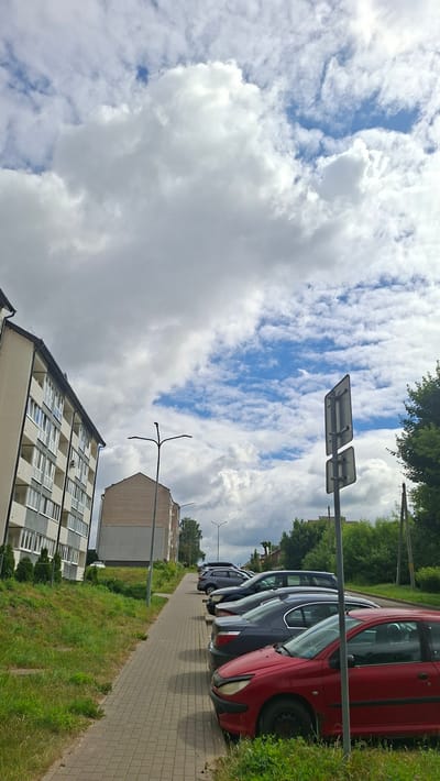 The media file displays a street scene in Smalyavichy, Belarus. The orientation of the image is such that the sky occupies the left side and the ground the right side. The sky is filled with a dramatic mix of white, fluffy cumulus clouds against a bright blue backdrop, suggesting it's a partly cloudy day, likely midday given the bright illumination.
On the right side of the frame, a row of parked cars is visible, lined up along a paved walkway. The cars are of various colors, including red, dark blue, and silver. The foreground features a patch of vibrant green grass bordering the walkway and the parked vehicles. Further back, a multi-story residential building with a light-colored facade and numerous windows is partially visible. Adjacent to it is another building, constructed with brickwork. A street lamp or light pole with a distinctive branching structure stands between the buildings. Two road signs, mounted side-by-side on a pole, are also prominent in the upper right quadrant of the image, facing away from the viewer. The presence of the residential buildings, cars, and paved walkway suggests an urban or suburban residential area. No people are visible in the scene. The overall atmosphere appears calm and ordinary, depicting a typical day in the depicted location.