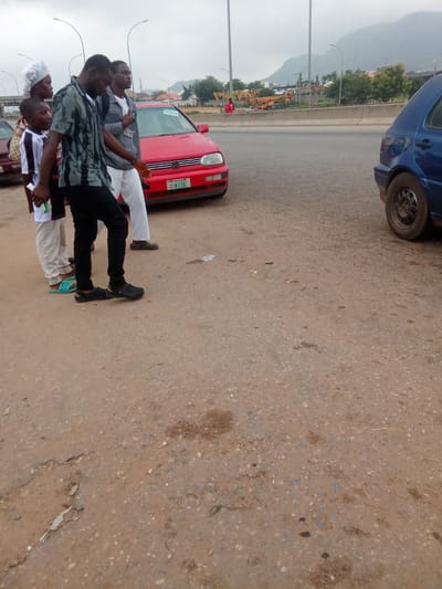 In Kagini, Nigeria, a group of four individuals, two men, a woman, and a boy, are walking on a dirt road next to a paved road where two cars, a red Volkswagen Golf and a blue hatchback, are parked. The red car is in the foreground on the left, while the blue car is in the background on the right. The individuals are casually dressed and appear to be on their way to or from an unspecified destination. The woman wears a white headscarf and a white shirt, while the boy is in a white and black shirt and light-colored pants. The two men are dressed in darker clothing. The environment is overcast, with a mountainous backdrop and some construction equipment visible in the distance.