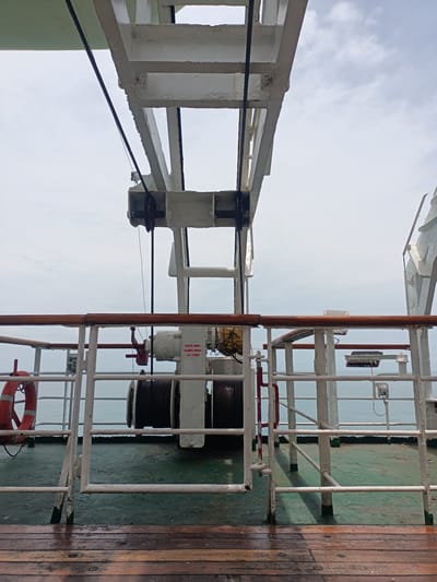 This image shows the deck of a ship, featuring safety railings, a winch, and an overhead crane structure. The deck is made of both wooden planks and painted green metal. A life preserver hangs on the railing to the left. The winch, a large cylindrical object with cable wound around it, is located centrally behind the railing. The date "10/2024" is visible on the winch's machinery. The overhead crane is a large white metal structure with cables running through pulleys. The background reveals the ocean and a cloudy sky. The location is assumed to be in Unknown, Malaysia, as per the prompt.