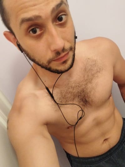 A shirtless man with a beard is facing the camera. He is wearing in-ear headphones. He appears to be indoors. There is no indication of the image being in Stafford, United Kingdom.