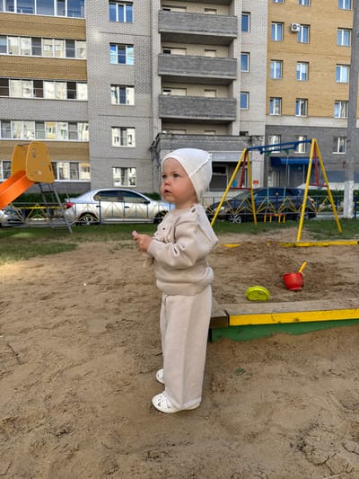 This is a full shot, taken outdoors, of a young child standing in a sandy area. The child is wearing a light-colored outfit consisting of a hat, a long-sleeved shirt, and pants. White shoes are visible on their feet. The child's gaze is directed slightly to the left, and their right hand is extended forward, holding a small pink object. In the background, there's a playground with a yellow and green border around the sandpit, and further back, a multi-story residential building with many windows. A dark car is parked next to the playground equipment. The sky is not visible, but the lighting suggests it's daytime. The scene appears to be a residential area, possibly a playground in a courtyard. The image is rotated 90 degrees counterclockwise.