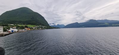 This media file captures a serene view of a fjord in Volda, Norway. The scene is dominated by a calm, dark body of water in the foreground, reflecting the overcast sky. In the mid-ground, a charming coastal village nestles at the foot of a verdant, tree-covered mountain. The village features a cluster of houses with colorful roofs, hinting at a residential area. Further along the coastline, industrial buildings with white and blue accents, along with some docks, suggest a small port or industrial zone. Red machinery and some parked vehicles are visible near the buildings.

The background is characterized by a series of majestic, mist-shrouded mountains, their slopes also covered in dense green forests. The sky is heavily clouded, with streaks of lighter clouds suggesting movement and a potentially cool, damp atmosphere. The overall mood is one of peaceful solitude and natural beauty. The time of day appears to be daytime, though the overcast conditions create a diffused light. The location is clearly identified as Volda, Norway, and the landscape features are typical of a Norwegian fjord. No people are explicitly visible, and no text is present in the image.