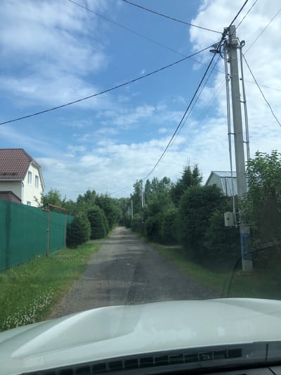 The image depicts a view from inside a vehicle looking out at a gravel road in the city of Veteran Stolitsy SNT, Russia. The road is lined with trees and bushes on both sides, with a tall concrete utility pole with numerous wires situated on the right side of the road. A green metal fence is visible on the left side of the road, with a white house with a brown roof behind it. The sky is blue with scattered white clouds. The hood of the vehicle is visible in the foreground.