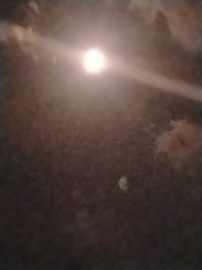 The image depicts a night sky with a bright, out-of-focus moon partially obscured by clouds. A hazy, luminous glow emanates from the moon, creating a soft light that illuminates the surrounding atmospheric conditions. The lower portion of the image shows a darker, less distinct area, possibly indicating ground level or a distant landscape. The overall impression is one of a serene, nocturnal scene, with the celestial body dominating the frame. The specific location in Almalyk, Uzbekistan, cannot be determined from the visual information provided.