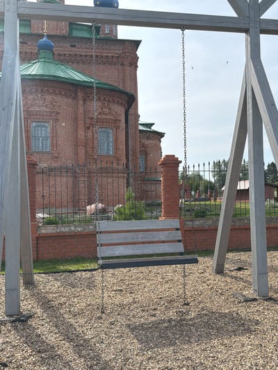 This is a photograph of a swing set in front of a brick church with green roofs and blue domes, likely located in Berezniki, Russia. The swing set has wooden planks for the seat and is suspended by metal chains from a wooden frame. The church is a prominent feature in the background, with intricate brickwork and arched windows. A metal fence separates the church grounds from the area where the swing set is located. The ground is covered in small gravel. The sky is overcast. There are no people visible in the image.