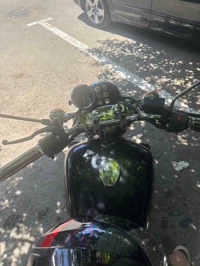 This is a high-angle, top-down shot of a black motorcycle parked on a paved surface, likely a street or parking lot, in Aytos, Bulgaria. Sunlight filters through overhead trees, casting dappled shadows across the motorcycle and ground. A portion of a dark-colored car is visible to the left of the motorcycle, with its silver wheel and tire prominent. The motorcycle features a glossy black fuel tank, chrome handlebars, and a circular speedometer or tachometer with a black face and white markings. Various controls and switches are visible on the handlebars. Part of a rider's leg, clad in a sandal, is visible on the right side of the frame, suggesting the motorcycle is occupied or has just been ridden. The scene appears to be set during daylight hours, with bright sunlight and clear shadows indicating fair weather. There is no discernible text in the image.