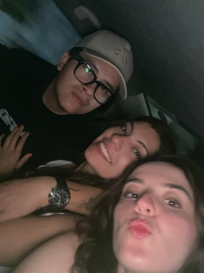 Three people are closely positioned together in what appears to be the interior of a vehicle. On the left, a person is wearing a tan-colored baseball cap with visible lettering, and thick-rimmed glasses. They are wearing a dark shirt. In the center is a person smiling with visible teeth. To the right, the third person is close to the camera, and they are pouting their lips. This person has a watch on their wrist. The scene appears dark, and the location is inferred to be within a vehicle, presumably in Brisas de Zicatela, Mexico.