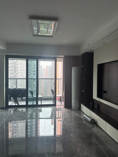 This image captures an interior view of a modern apartment, likely in Shunde District, China, showcasing a living area with a large glass door leading to a balcony. The room features polished marble flooring, which reflects the exterior buildings and the interior ceiling light. To the left, through the glass door, a balcony is visible with two chairs, a clothes drying rack with dark fabric draped over it, and a view of numerous high-rise residential buildings. Inside, a tall, white air conditioning unit stands near a dark curtain. The right side of the room is dominated by a built-in entertainment unit with dark shelving and white cabinets. A large dark television screen is mounted on the unit. On the floor in front of the entertainment unit, a rolled-up mat is visible. The ceiling has a large, square, modern light fixture.