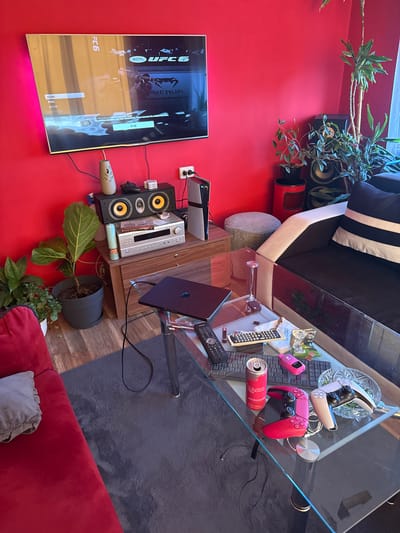 The image depicts a living room interior with a vibrant red wall, suggesting a modern or eccentric decor choice. Prominently featured is a large flat-screen television displaying the UFC 6 video game menu, with a message indicating "The wireless controllers have been disconnected." Below the television, a black speaker system sits atop a wooden cabinet. On the cabinet, various items are placed, including a silver stereo receiver, a potted fiddle-leaf fig plant, and a PlayStation 5 console.

In the foreground, a glass-top coffee table holds a collection of items. These include a laptop, a black remote control, a pink gaming controller, a silver gaming controller, and a can of "PEACH RUSH" energy drink. A variety of other small objects, such as keys, a bag of snacks, and what appears to be a USB drive, are scattered across the table. A red sofa is visible to the left of the frame, partially covered by a grey throw pillow. To the right, a dark sofa with striped cushions is positioned, and further back, several potted plants add a touch of greenery to the room. The overall environment suggests a casual setting, likely a home entertainment area. There are no individuals visible in the image. The location is identified as Kostinbrod, Bulgaria.