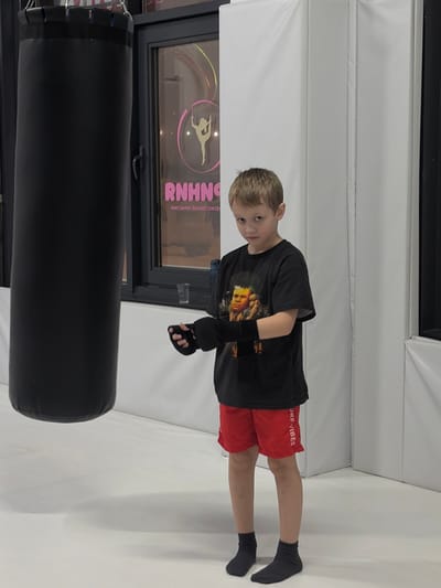 An interior image taken in Novosaratovka, Russia, depicts a young male subject standing in a combat sports or fitness facility. The subject, a boy estimated to be between five and eight years old, is positioned towards the right of the frame, looking directly at the camera with a neutral expression. He wears a black t-shirt featuring a graphic of an older male, red athletic shorts with "OFF VIBES" printed vertically on the left leg, and black socks. Black fingerless hand wraps are present on both of his hands. To the left of the subject, a large black cylindrical punching bag hangs from an unseen fixture above, partially visible. Behind the punching bag and the subject, a dark-framed window reveals a pink neon-style sign inside what appears to be an adjacent space or reflection. The sign features a stylized female figure in a dynamic pose within a circle, above the word "RNHNGA" and mirrored Russian text below, which translates to "CONTACT PHONE NUMBER." A small clear glass and a blue water bottle are visible on a ledge beneath the window. The floor is covered with a light-colored mat, and white padded walls extend along the right side and corner of the background.