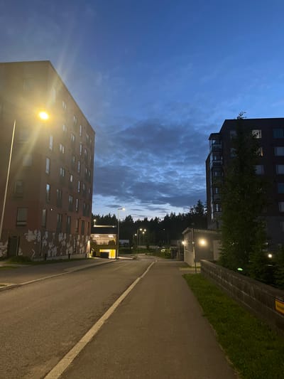 The image depicts a street scene at dusk in Vantaa, Finland. The sky is a deep blue with scattered, dark clouds, suggesting the transition from daylight to night. On the left side of the frame is a tall, multi-story residential building with a red facade. Its windows are dark, and a bright streetlamp illuminates its side, casting lens flares. The lower section of this building features abstract white murals.

To the right of the first building, a paved road curves gently away from the viewer. Adjacent to the road is a pedestrian walkway. Further down the road, a yellow structure, possibly a parking garage entrance or a building amenity, is illuminated by yellow lights.

On the right side of the image, another residential building, this one with a dark facade, is visible. It also has multiple stories and visible windows, some of which are lit from within. A large, leafy tree stands in front of this building. A low, dark wall runs along the edge of the walkway, with a strip of green grass behind it. Streetlights are visible in the distance, lining the road, and a dense line of trees forms the backdrop at the far end of the scene. The overall impression is one of a quiet, residential urban environment at the end of the day.