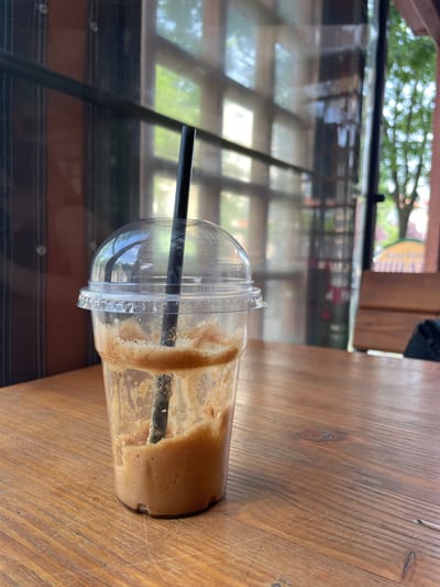 The image, captured in Nova Zagora, Bulgaria, features a partially consumed frothy beverage in a clear plastic cup. The cup, fitted with a dome lid and containing a black straw, lies on its side on a warm-toned wooden table. The beverage, a light brown color resembling iced coffee or a similar drink, exhibits foam and residue along the inside walls of the cup, indicating it has been partially drunk.
The setting appears to be an indoor space, likely a cafe or restaurant, characterized by the wooden table and large windows. The windows prominently display reflections of an outdoor environment, including blurred green foliage from trees and bright natural light, suggesting it is daytime. Distant architectural elements and a highly blurred yellow sign are also faintly visible through the reflections.
Notable details include fine scratches and wood grain visible on the table surface. On the rim of the plastic cup's lid, faint molded text such as "PET" and "RECYCLE" can be discerned. No people are visible in the frame, and the scene conveys a calm, still atmosphere.