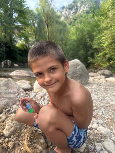 A young boy with short, dark hair and brown eyes smiles at the camera while crouching on a rocky riverbank. He is shirtless and wearing blue patterned swim trunks. In his right hand, he holds a small, clear plastic bottle with a green cap, which appears to contain some small objects. The boy's posture suggests he is engaged in an activity, possibly examining or collecting something from the river.
The setting is outdoors, with lush green trees and foliage visible in the background. Large, grey rocks are scattered around the riverbank, and a body of water, likely a river, is present to the left of the boy. The sunlight suggests it is daytime, and the overall atmosphere appears relaxed and natural. The location is identified as Saint-Claude, France. No visible text is present in the image.