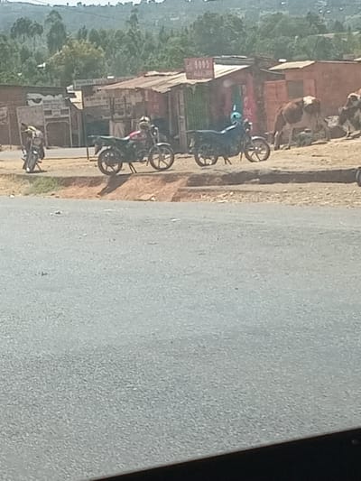 An outdoor scene in Kipkarren, Kenya, depicts a bustling roadside area under bright daylight. The image, rotated counter-clockwise, shows a grey asphalt road on the right, with a dirt and gravel shoulder bordering it. To the left of the road are several simple, informal structures, likely shops or stalls, constructed from corrugated metal and wood, featuring flat or gently sloped roofs.

Several motorcycles, commonly known as "boda-bodas," are prominent subjects. At least three motorcycles are clearly visible in the midground, parked or standing near the buildings, with one further back. At least three individuals are present, primarily associated with the motorcycles; one person near a blue motorcycle is wearing a light blue or teal top. Two cows can be seen standing near one of the buildings in the background.

The setting suggests a rural or semi-urban commercial hub. In the distant background, lush green hills and trees indicate a natural, possibly elevated, landscape. The bright light suggests it is daytime with sunny weather.

Visible text on the signs of the buildings includes partially discernible words like "GOODS," "100," "NORTH," "MARKET," and "WHOLESALE," confirming the presence of commercial establishments. The overall activity appears to be typical daily street life, with people and motorcycles gathering near these roadside businesses.