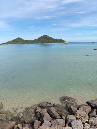 The image presents a seascape with a clear, turquoise sea in the foreground, transitioning to a brighter blue under a partly cloudy sky. In the distance is a lush, green island with two prominent hills. The shoreline in the immediate foreground is composed of dark, rocky formations. The water is clear enough to reveal the sandy seabed near the rocks. The location is suggested to be Layeun, Indonesia. There is no explicit evidence of human presence or interaction in the immediate scene, beyond the implication that the image was taken from a human perspective on the shoreline.