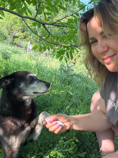 An outdoor scene in Leningradskaya, Russia, depicts an adult female interacting with a dog under a leafy tree canopy during daylight. The woman, positioned on the right, has blonde hair and is partially visible from the right side, looking towards the dog with a slight smile. She wears a dark grey t-shirt with partial Cyrillic text visible on the shoulder ("КОСМС"). Her right hand, featuring bright pink manicured nails, is holding the right paw of a medium-sized dog positioned on the left. The dog has black fur with a grey and white muzzle and chest, indicative of age, and is looking towards the woman's direction. Both subjects are on lush green ground covered in various grasses and plants. Overhead, a prominent tree branch with green leaves casts shadows, creating a dappled light effect. In the distant background, through the foliage, a wire fence and vague structures are visible.