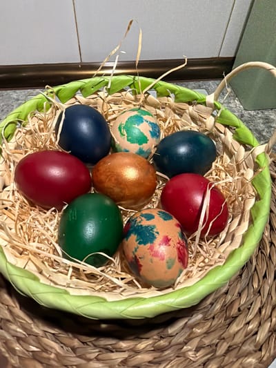 A close-up captures a green braided basket filled with straw and eight Easter eggs. The eggs are of varying colors: red, blue, green, bronze, and patterned with combinations of green, red, and blue. The basket sits on a woven mat. The scene appears indoors. The photograph was taken in Azuqueca de Henares, Spain.