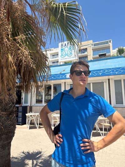 A young man, wearing sunglasses and a blue polo shirt, poses with his hands on his hips. He is standing outdoors in front of a building with a blue awning decorated with fish motifs. Behind him, a palm tree with dry fronds is visible, along with outdoor seating and tables. The building appears to be a restaurant, as indicated by the text "BAM! RESTAURANT & DELICIOUS" on its facade. The scene is bathed in bright sunlight, suggesting it's daytime. The man's pose and expression convey a sense of casual confidence. The location cues, such as the architecture and the palm tree, are consistent with a Mediterranean setting, likely in Gżira, Malta.