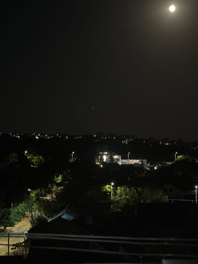 This is a night-time, high-angle photograph of a residential area in Cahul, Moldova. The sky is dark with a bright full moon visible in the upper right quadrant. A diffused glow from artificial lighting illuminates the scene below. The landscape is characterized by dark silhouettes of trees, rooftops, and the gentle slopes of hills. Numerous streetlights and lights from buildings are scattered throughout the image, creating points of illumination against the dark urban sprawl. In the foreground, horizontal lines suggest a balcony railing or fence. The overall impression is of a quiet, illuminated town at night, viewed from a distance.
