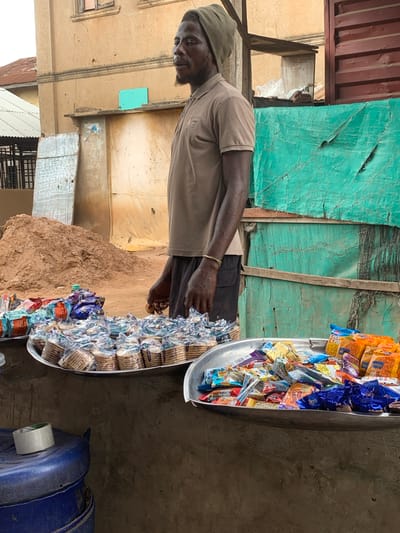 A man stands behind a stall displaying various packaged snacks and biscuits in Shagamu, Nigeria. The man wears a brown short-sleeved polo shirt and dark shorts, with a green head covering. He is positioned to the right of the frame, looking towards the left. In front of him are two large, round metal platters filled with an assortment of individually packaged snacks. One platter, closer to the viewer, contains brightly colored snack bags. The platter behind it is filled with small, stacked packages of biscuits. To the left of the platters, a blue plastic container is visible, with a roll of tape on top. The stall is set up against a building with textured walls and a corrugated metal roof. Behind the stall, a large mound of reddish-brown sand is present. The background also features green tarpaulins stretched across wooden frames. The scene appears to be an outdoor market stall.