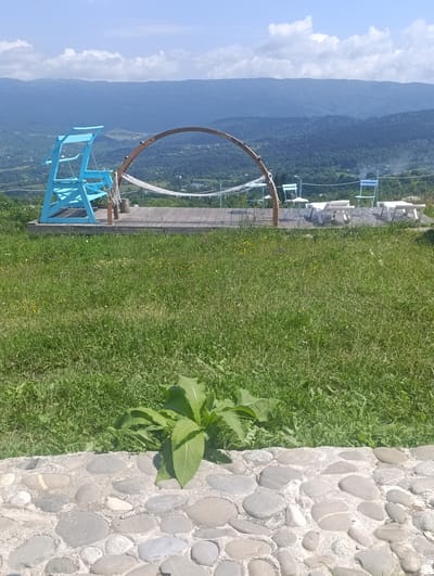 The image depicts an outdoor recreational area on a hillside, overlooking a valley with rolling, tree-covered mountains in the distance. In the foreground, a stone pathway lines a lush green lawn dotted with wildflowers.
On a wooden deck situated on the slope, there are several pieces of outdoor furniture. A bright blue, multi-level swing set is positioned on the left. Adjacent to it, a rustic wooden archway supports a white hammock. Further to the right, white lounge chairs and small tables are arranged. A few pale blue folding chairs are also visible. The environment suggests a peaceful setting, likely a tourist or leisure spot. The location is identified as Nikortsminda, Georgia.