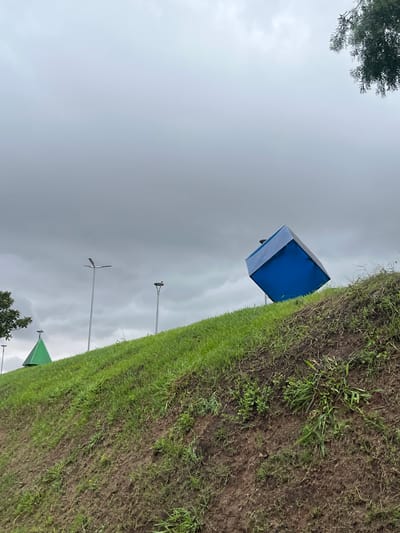 The image presents an outdoor landscape in Avellaneda, Argentina, under an overcast sky. A prominent feature is a grassy and muddy incline occupying the right side of the frame, contrasting with the expansive grey sky on the left.
Perched atop the grassy slope, slightly off-center, is a blue and light-colored portable toilet or temporary booth, appearing to face towards the left. The ground is a patchwork of green grass and dark, exposed soil, suggesting damp conditions or an area that has seen some foot traffic. In the mid-ground, against the dull, cloudy sky, several tall poles resembling streetlights or utility poles are visible. One distant pole on the right seems to have a small bird resting on its top.
Dark green foliage from a tree overhangs into the top-left corner of the frame, while a glimpse of a green, triangular structure, possibly a tent or roof, and more foliage can be seen in the bottom-right corner. The scene depicts a quiet, static moment with no people or specific activities taking place. The heavy cloud cover indicates a daytime setting under dull weather conditions. No text is visible in the image.
